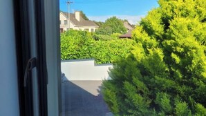A view from a window overlooking a street with a hedge and a white wall.
