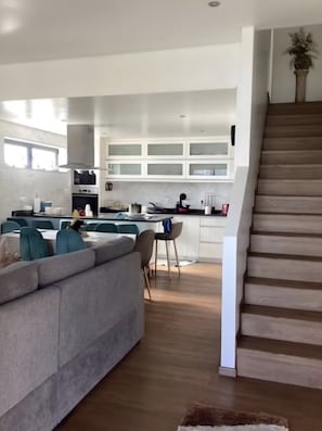 Una cocina moderna con armarios blancos, un comedor con sofá gris y una escalera que sube a un nivel superior.
