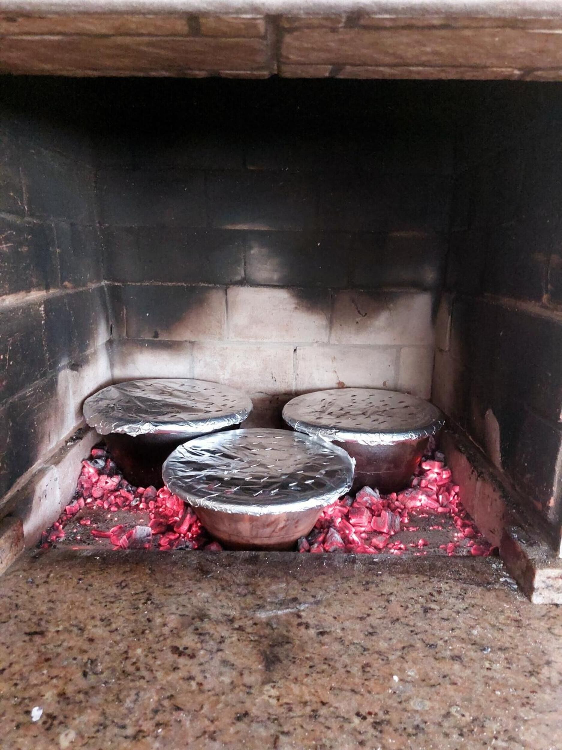 A brick oven with four metal containers on a bed of glowing coals.