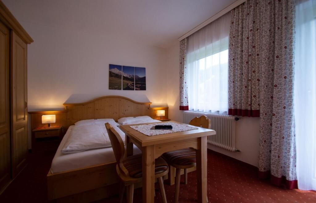 A room with a bed, two bedside tables with lamps, a wooden table with chairs, a window with curtains, and a radiator.