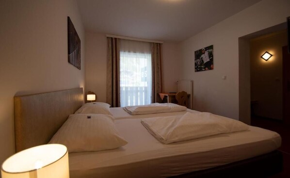 A hotel room with two beds, a desk, a chair, a window with curtains, and a wall-mounted light.