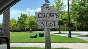 A wooden sign with the text 'The CROW'S NEST' in a residential area with a sidewalk, trees, and a clear sky.