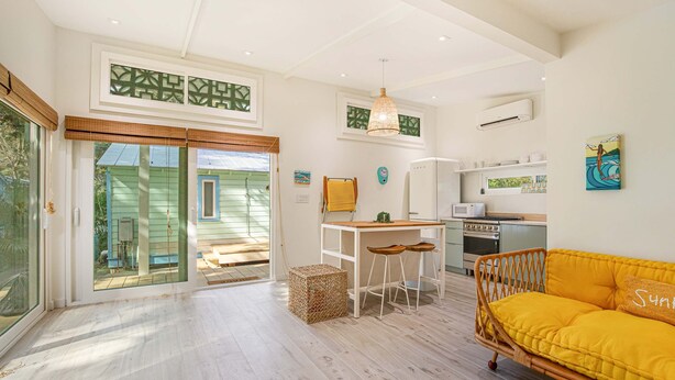 A bright, open-plan living area with a kitchen, a yellow sofa, and a dining table with stools.