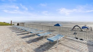 Eine Strandlounge mit Liegestühlen und Sonnenschirmen, einem Metallgeländer und Blick auf Strand und Meer.