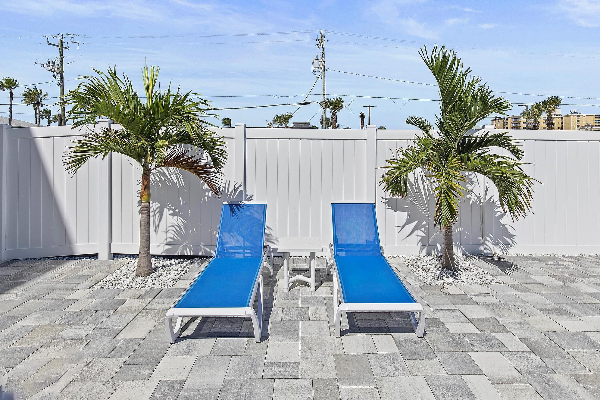 Deux fauteuils de jardin bleus avec des structures blanches, une clôture blanche et des palmiers dans un espace extérieur ensoleillé.