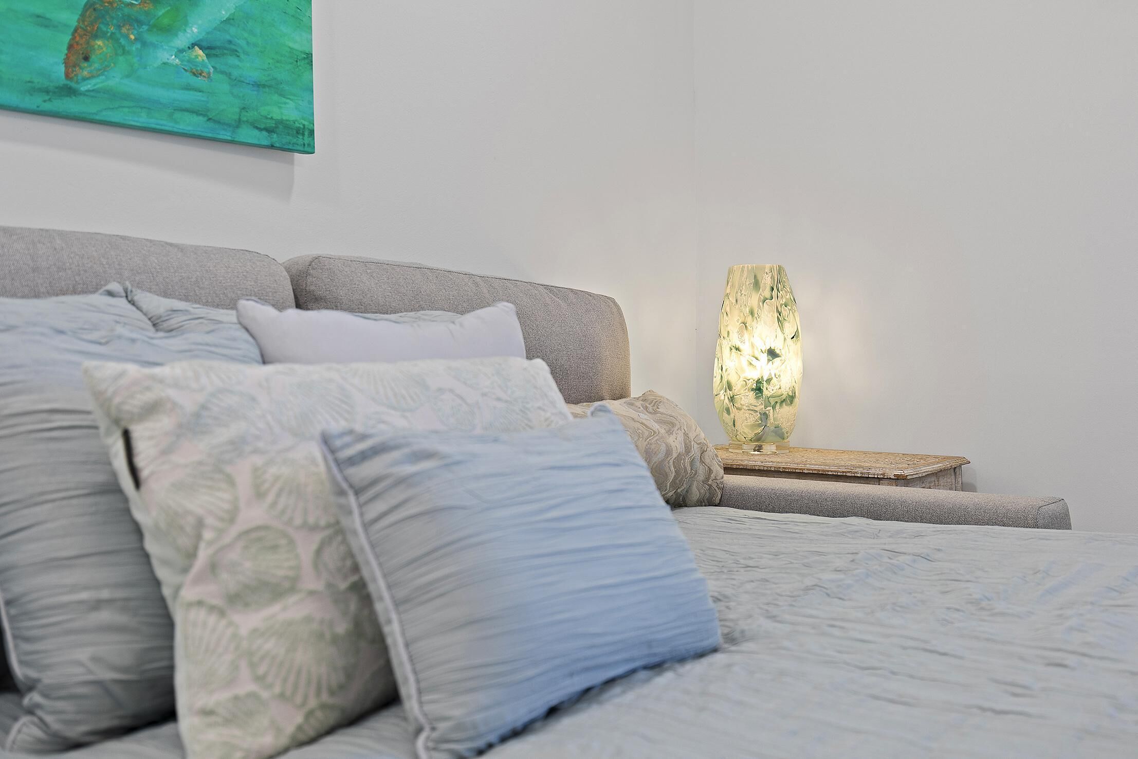A neatly made bed with a grey headboard, a bedside table with a lamp, and a painting on the wall.