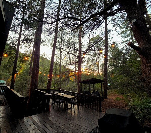 A wooden deck with tables and chairs, surrounded by tall trees and string lights. - Lavender Cottage at Kohl's Ranch- Across from Tonto Creek (Payson)