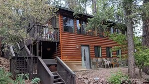 A log cabin with a wooden staircase leading to a deck, surrounded by trees and a stone wall.