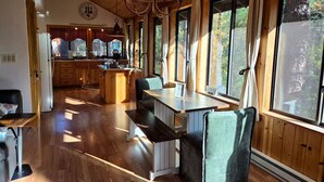 A cozy cabin interior with a wooden dining area, large windows, and a kitchen in the background.