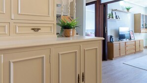 Una cocina con armarios de color crema, una escalera de madera y vistas a una sala de estar con televisor y plantas.