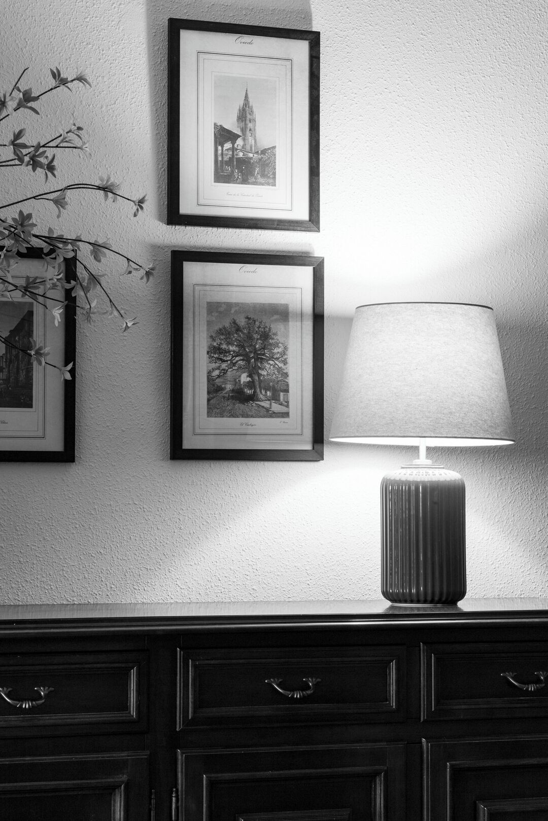 A black and white photo of a wall with three framed pictures and a lamp.