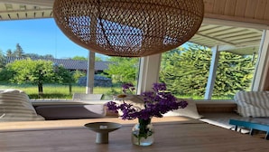 A wooden table with a clear view of a garden through large windows, a wicker hanging light fixture, and a vase with purple flowers. - 5 Star Holiday Home in Hundested (Hundested)