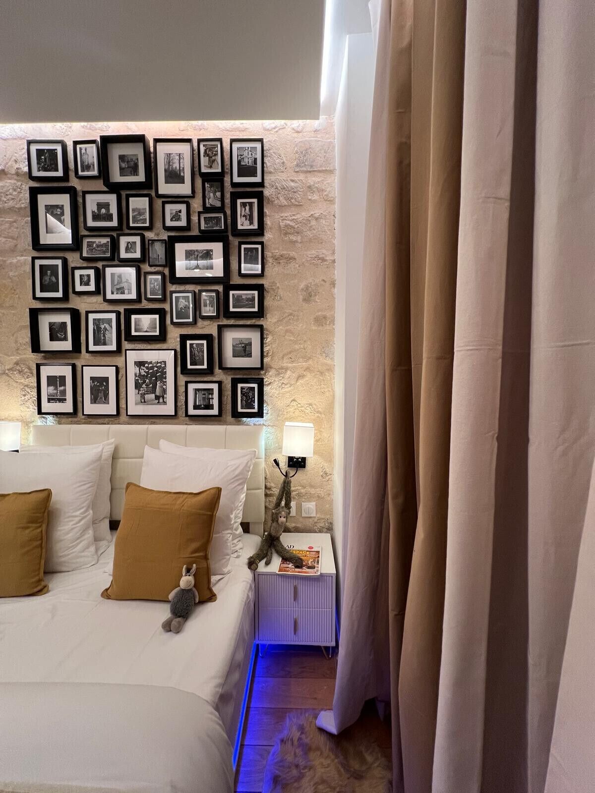 A hotel room with a bed, a wall of framed photos, a nightstand with a lamp, and a curtain.