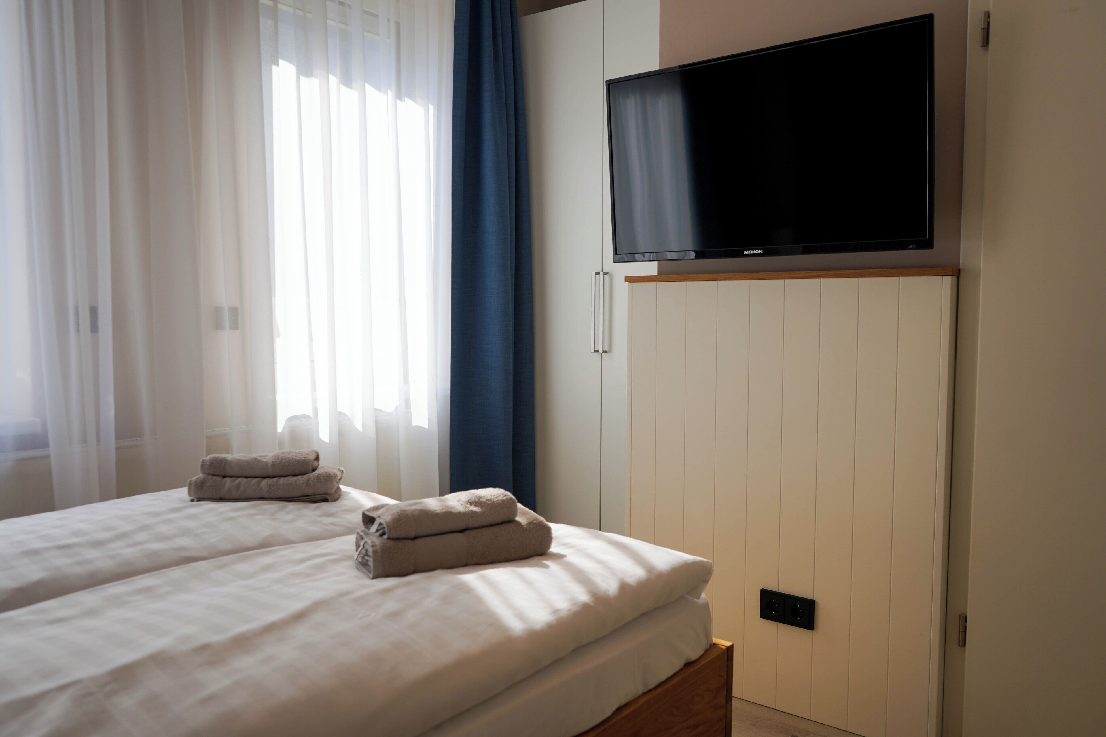 A hotel room with a bed, two towels, a television on the wall, and a window with curtains.