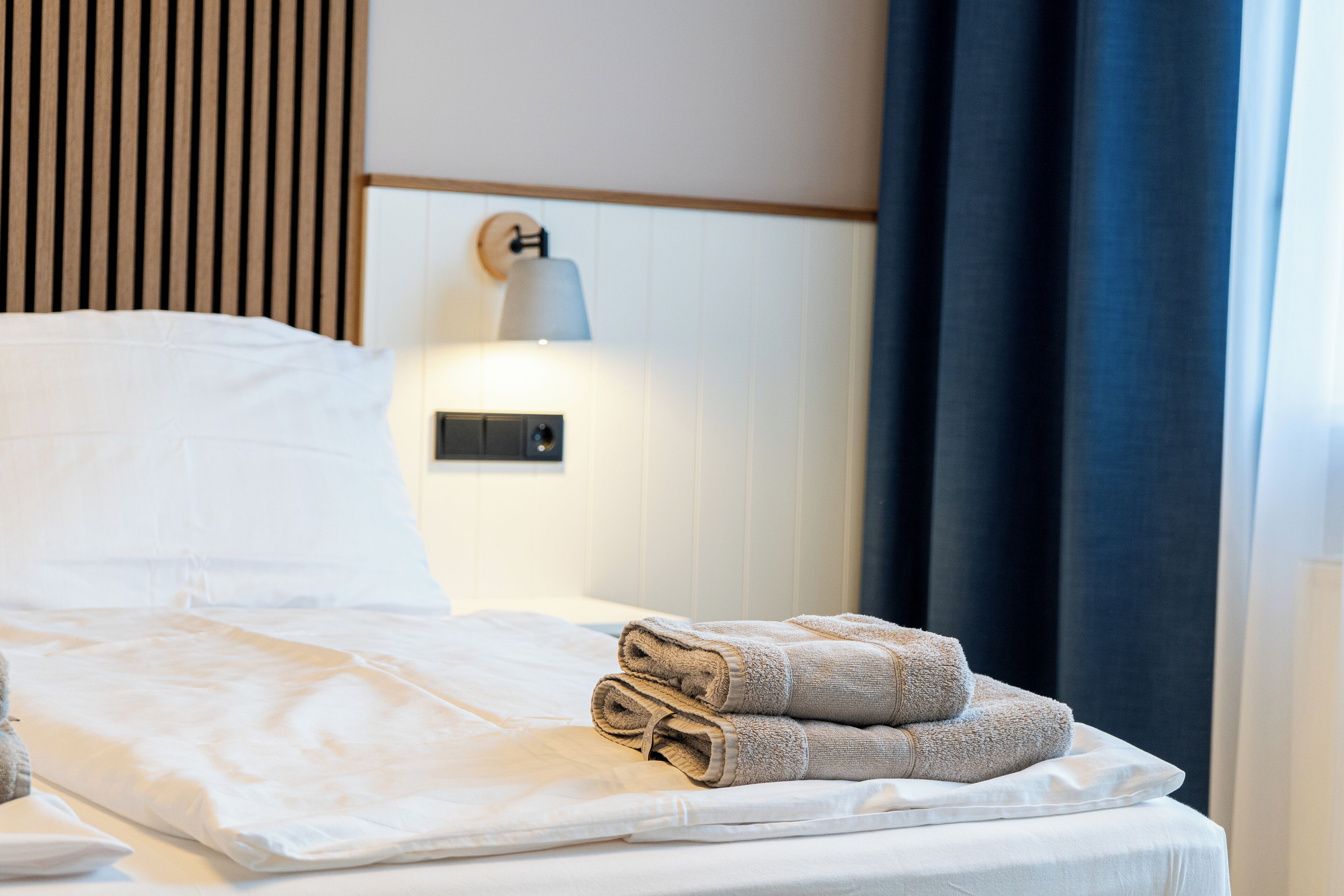 A neatly made bed with white linens and two rolled towels on the side.