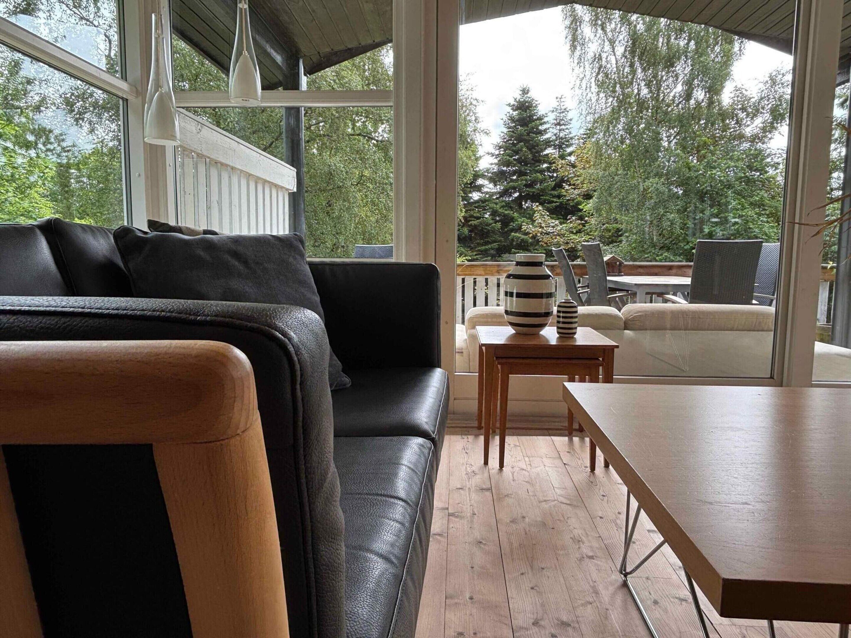 A modern living room with a black leather sofa, a wooden table, and a view of the outdoors through large windows.