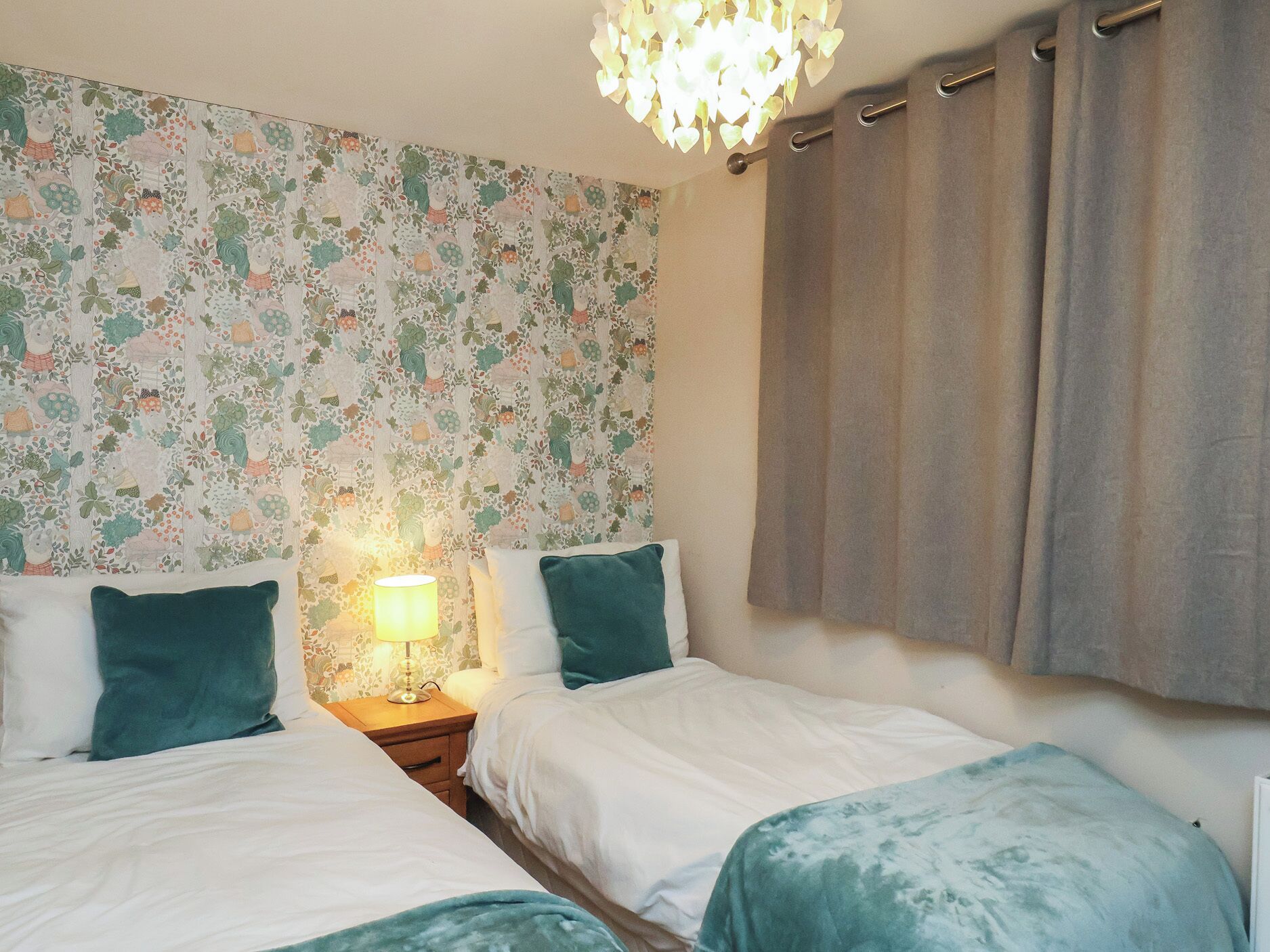 A bedroom with two beds, a nightstand with a lamp, floral wallpaper, and a chandelier.