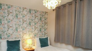 A bedroom with two beds, a nightstand with a lamp, floral wallpaper, and a chandelier. - Farriers Yard (Whitby)