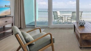 Una sala de estar con un sofá, una mesa de centro de madera y una silla. A la izquierda, hay un televisor sobre un mueble bajo. La habitación tiene un amplio ventanal corredizo que da a un balcón con vistas a la playa y al océano.