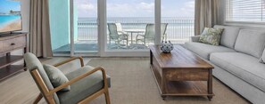 Un salon avec un canapé, une table basse en bois et un fauteuil. Une télévision est posée sur un meuble à gauche. La pièce est dotée d’une grande porte-fenêtre coulissante qui donne sur un balcon offrant une vue sur la plage et l’océan.