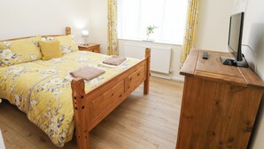 Ein Schlafzimmer mit einem hölzernen Bett, einem Nachttisch, einem kleinen Tisch daneben, einem Fenster mit Vorhängen, einer Heizung und einem Fernseher auf einem Holzgestell.