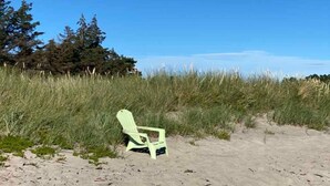 A beach scene with a single green chair in the foreground, surrounded by tall grass and trees in the background.