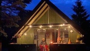 A small, green-roofed cabin with a porch, a fire pit, and outdoor seating.