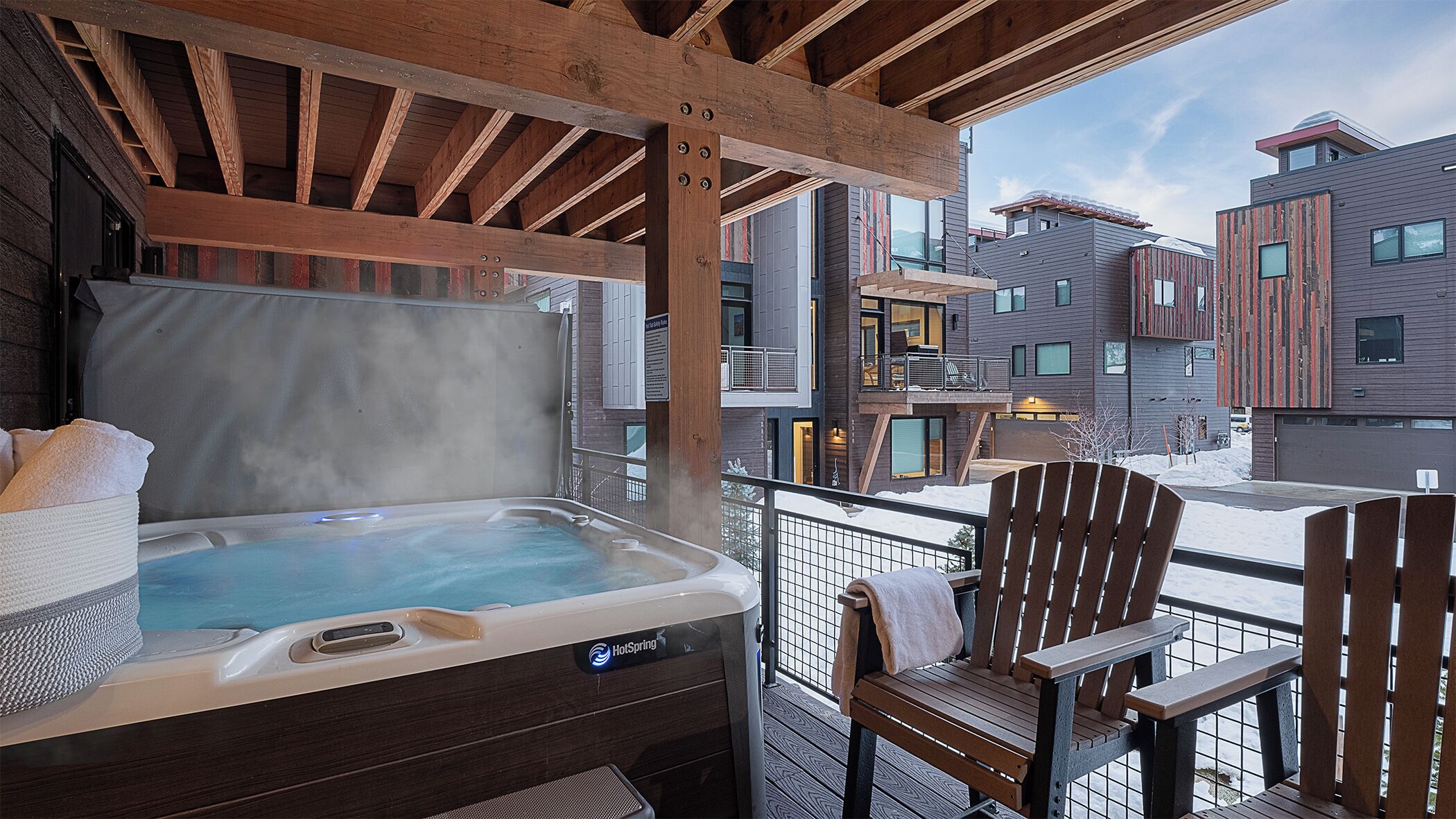 Un jacuzzi en un balcón con sillas de madera, con vistas a un complejo de edificios moderno.