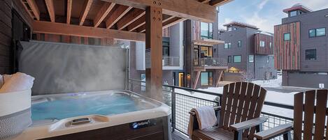 Un jacuzzi en un balcón con sillas de madera, con vistas a un complejo de edificios moderno.