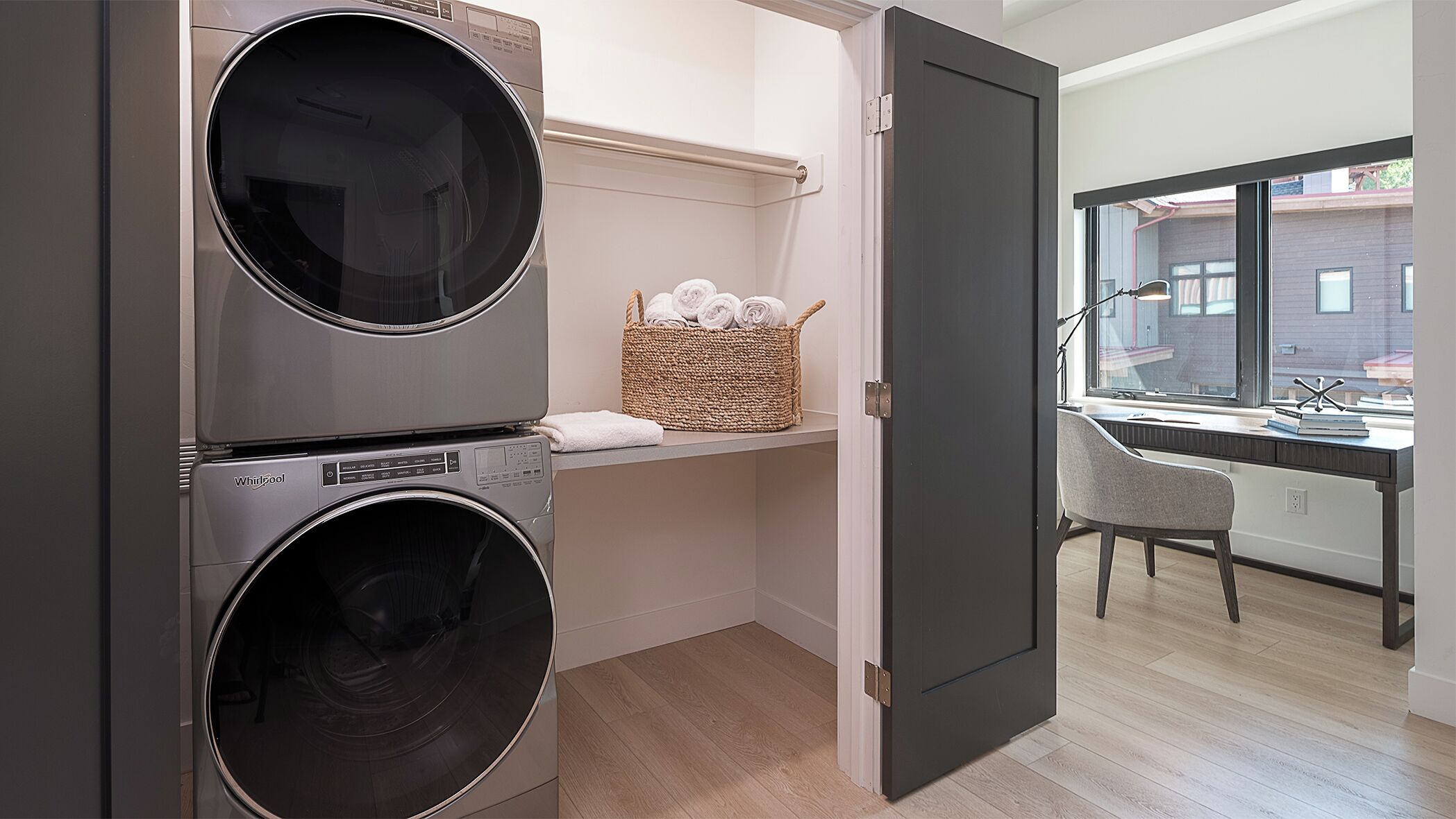 A laundry room with two stacked front-loading washers and dryers, a wicker basket with folded towels, and a view of a building through a window.