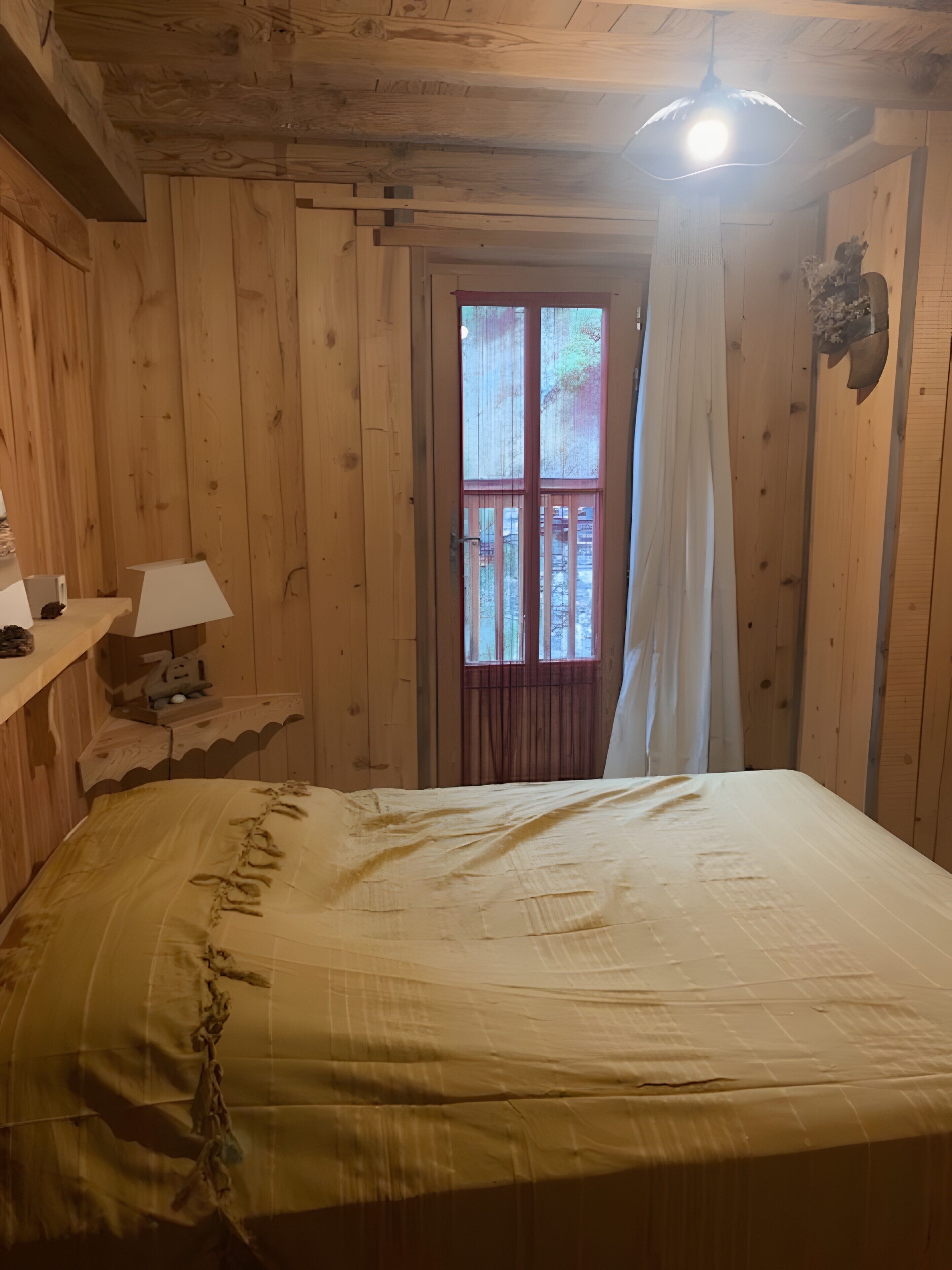 Ein Zimmer mit Holzboden, einem Bett, einem Fenster mit Vorhängen, einer Lampe und einem Regal.