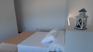 Ein Einzelbett mit weißer Bettdecke und einer beigen Tagesdecke, ein Nachttisch aus Holz und eine wandmontierte Leuchte.