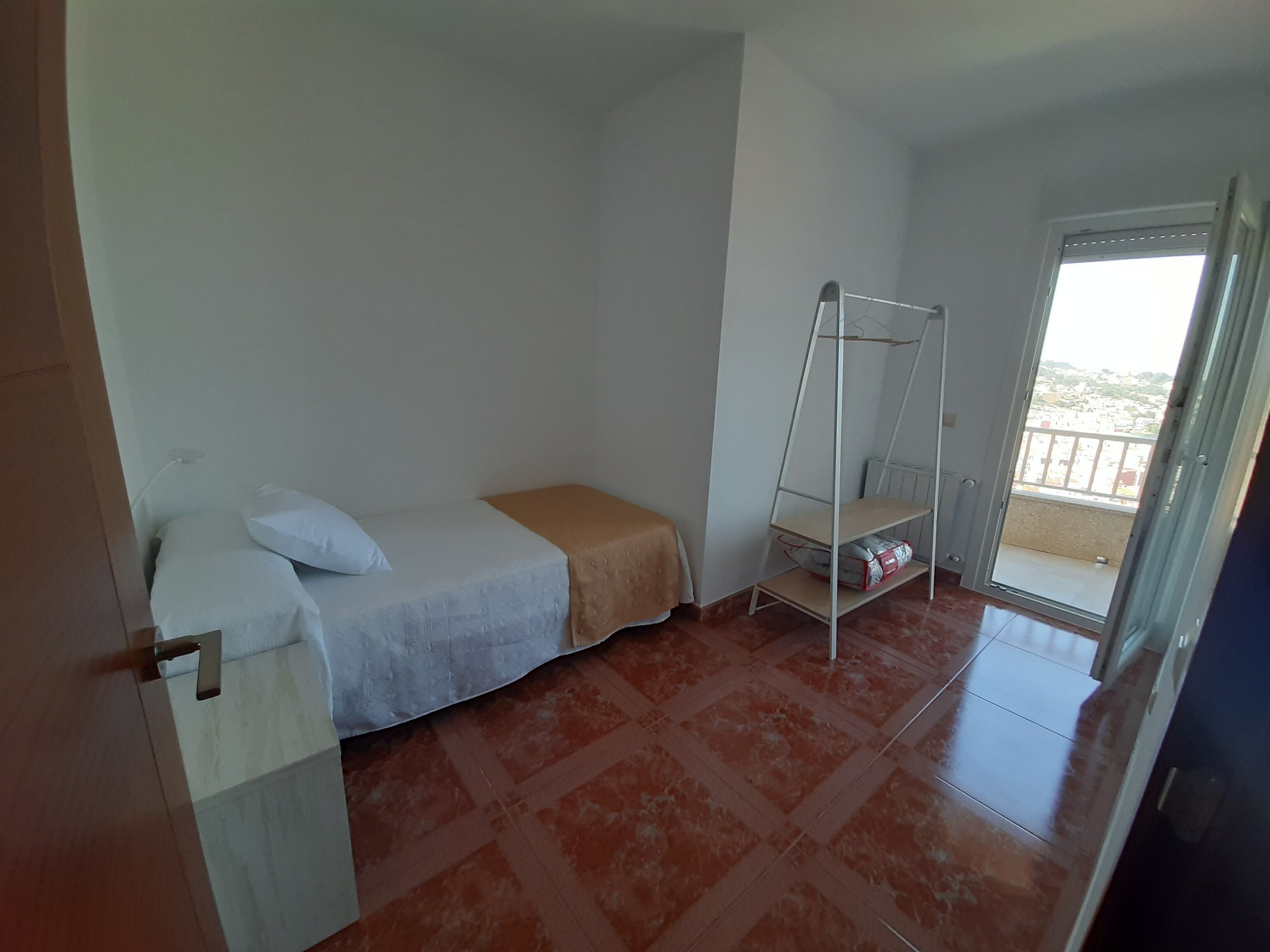 Habitació petita i senzilla amb un llit, una estantària de metall i vistes per una porta.