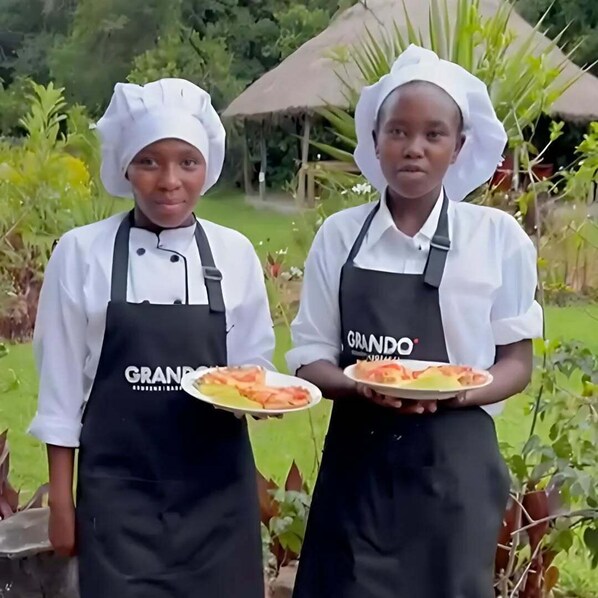 Two individuals in chef attire holding plates of food, standing outdoors with a thatched-roof structure and greenery in the background. - Oreteti Mara Lodge (Lolgorien)