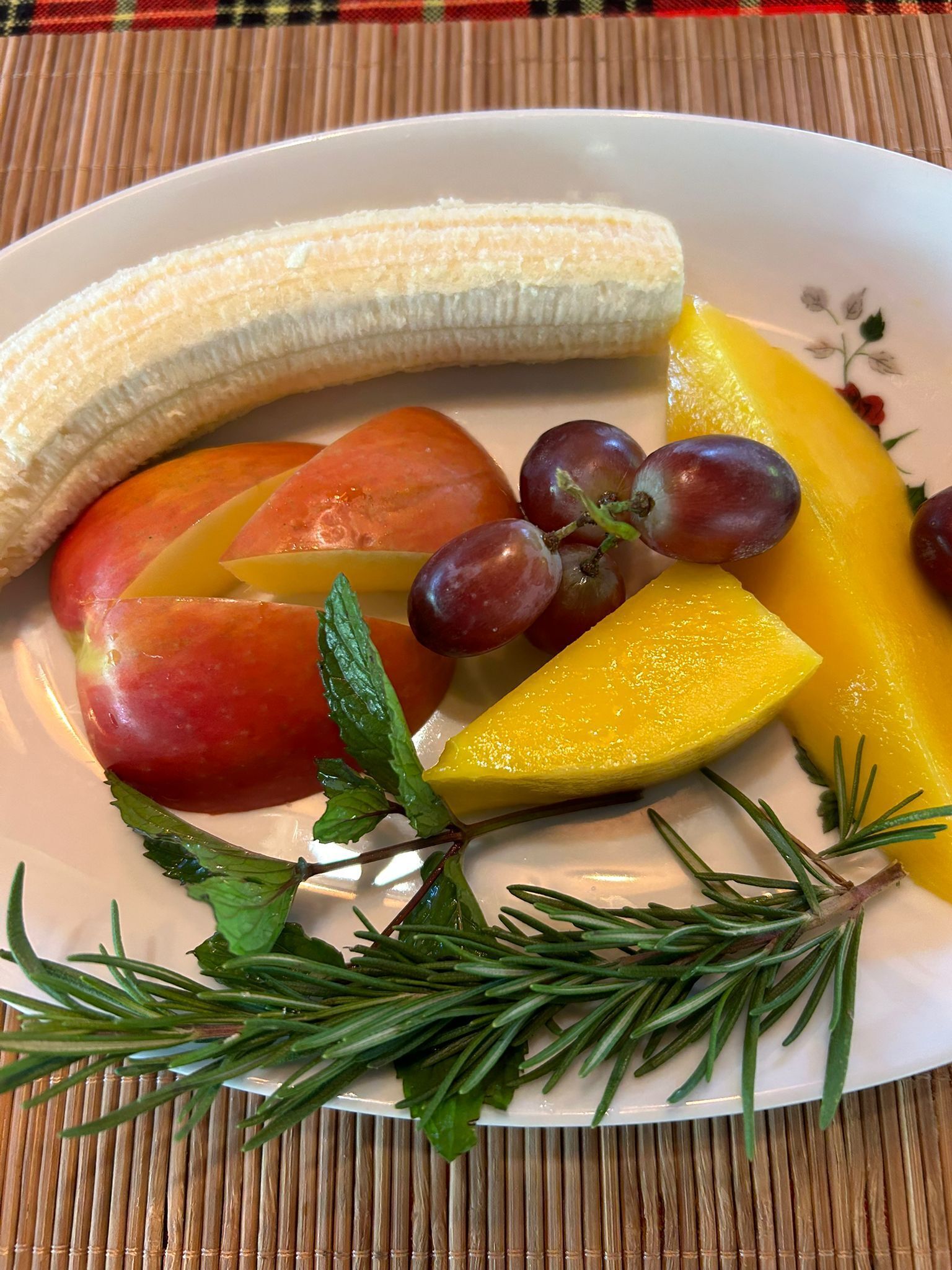 Prato com frutas fatiadas, incluindo banana, manga e pêssego, decorado com alecrim.