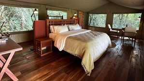 A spacious tent with a wooden floor, a bed with a white bedspread, a wooden nightstand, a chair, and a table.