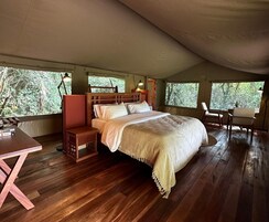 Una carpa espaciosa con piso de madera, una cama con cubrecama blanco, una mesita de noche de madera, una silla y una mesa.