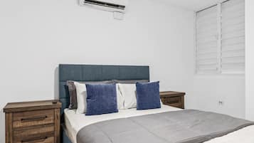 Ein Schlafzimmer mit einem Bett, zwei Nachttischen, einem Schreibtisch, einer Klimaanlage und einem Fenster mit Jalousien.