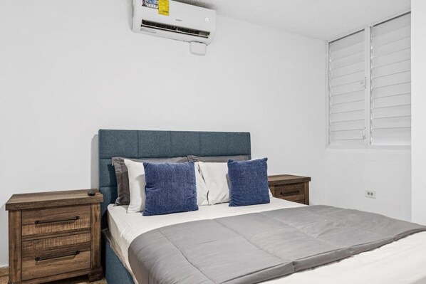A bedroom with a bed, two bedside tables, a nightstand, an air conditioning unit, and a window with blinds.