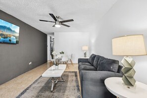 Ein modernes Wohnzimmer mit einem grauen Sofa, einem Couchtisch, einem an der Wand befestigten Fernseher und einem Deckenventilator.