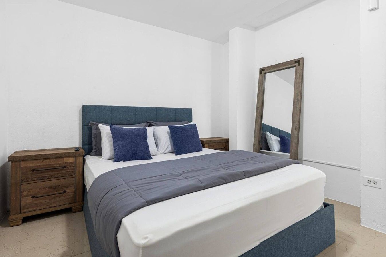 Een slaapkamer met een bed, een houten nachtkastje, een spiegel en een witte muur.