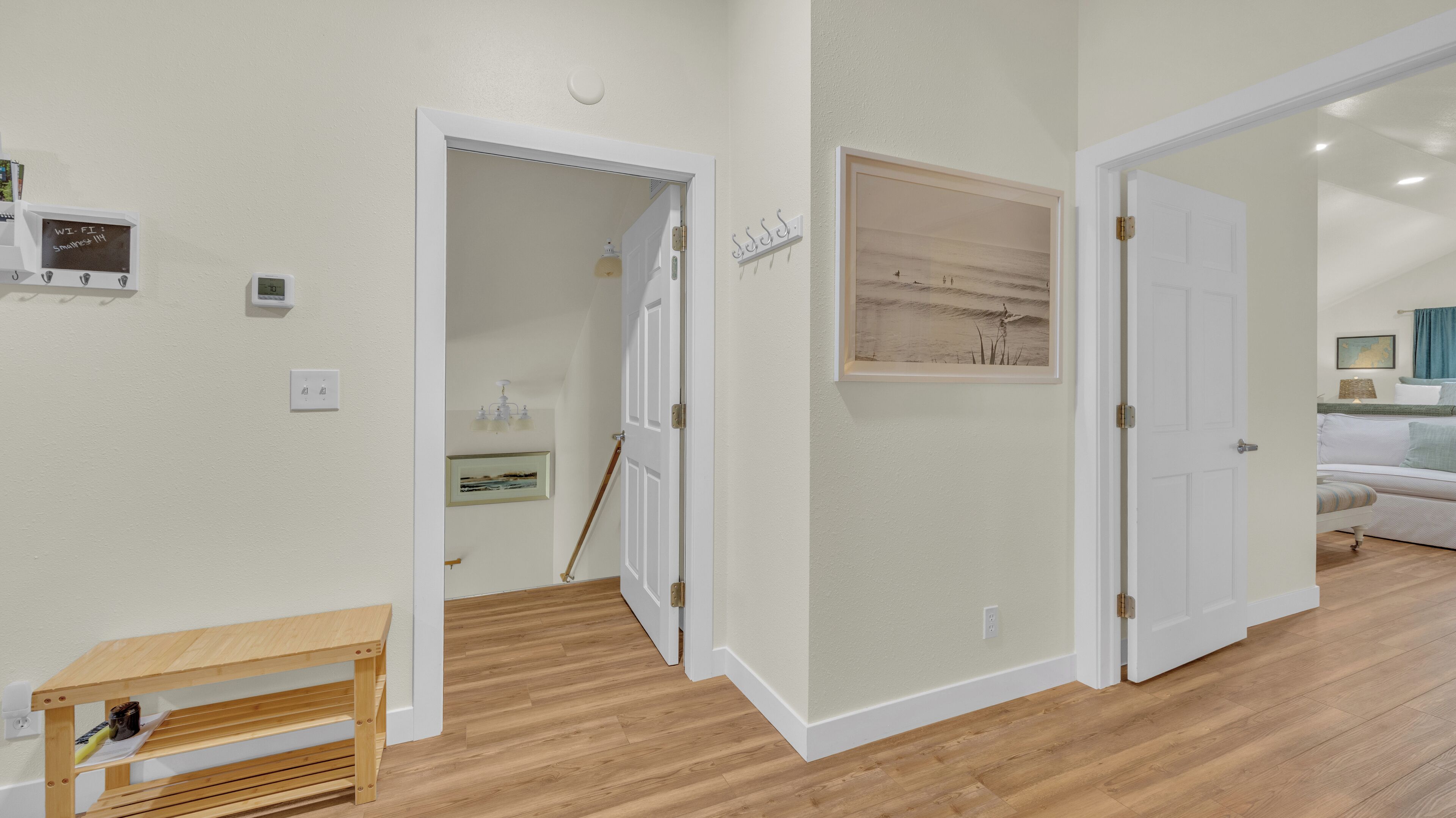 Un pasillo con piso de madera, paredes blancas y un banco. Hay puertas que dan a otras habitaciones, una pequeña mesa con una botella, y una lámpara fijada a la pared.