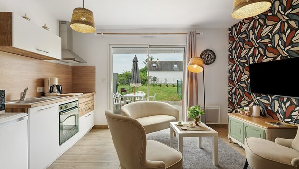 Une cuisine moderne équipée d’une machine à café, un coin repas avec une table et des chaises, et un salon avec un téléviseur et un mur décoré de motifs.