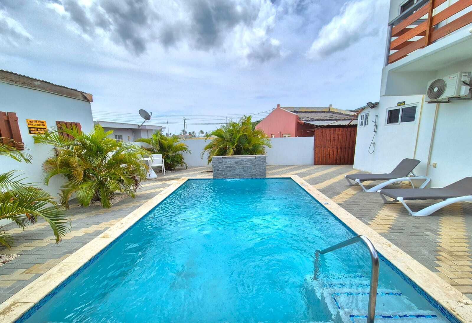 A rectangular swimming pool with a tiled deck, surrounded by palm trees and a red-roofed building.