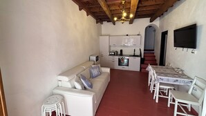 Een ruimtelijk ingerichte leefruimte met een bank, eettafel, stoelen, een kitchenette en een aan de muur bevestigde televisie.