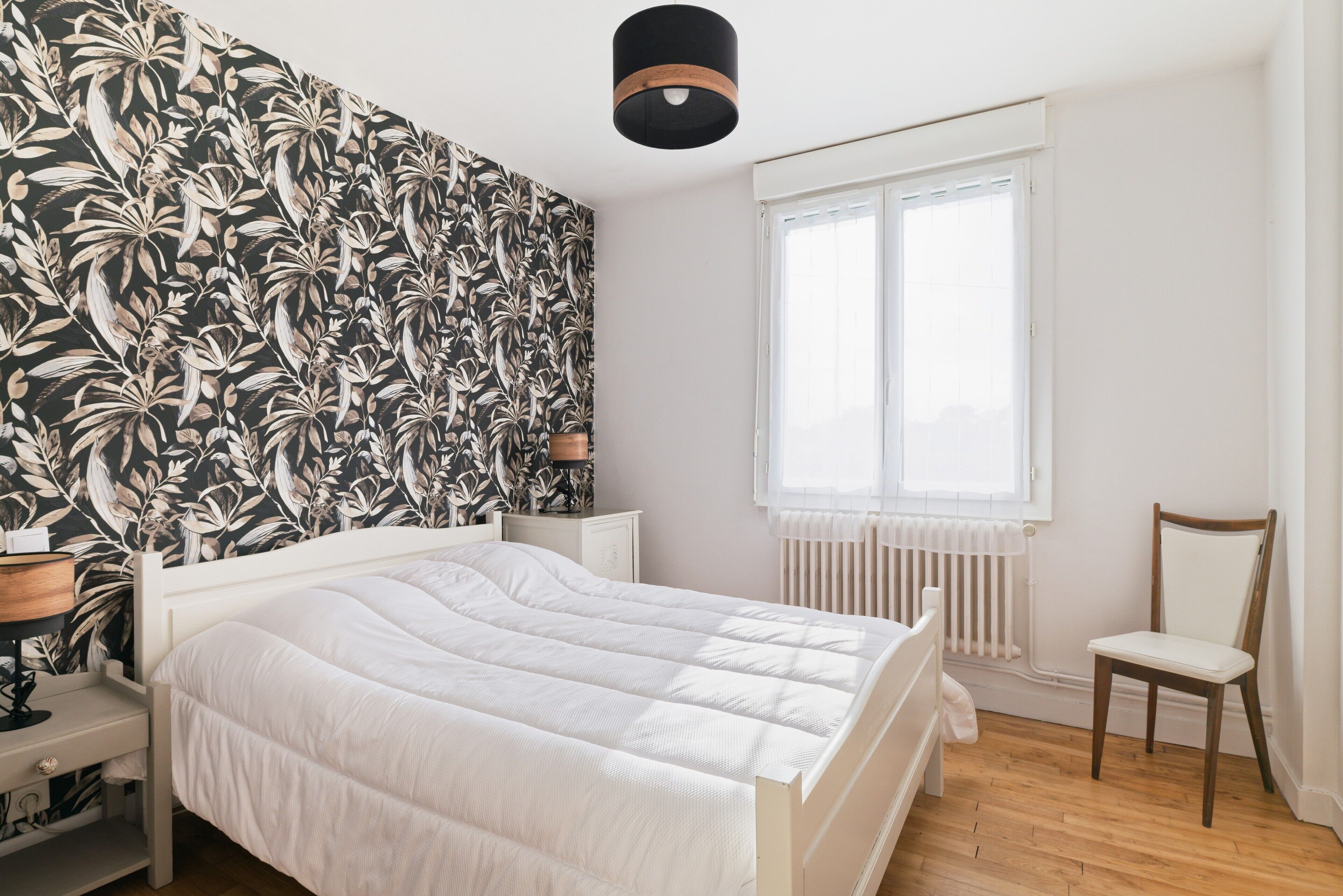 Een slaapkamer met een bed, een stoel, een nachtkastje, een lamp, een radiator en een raam.