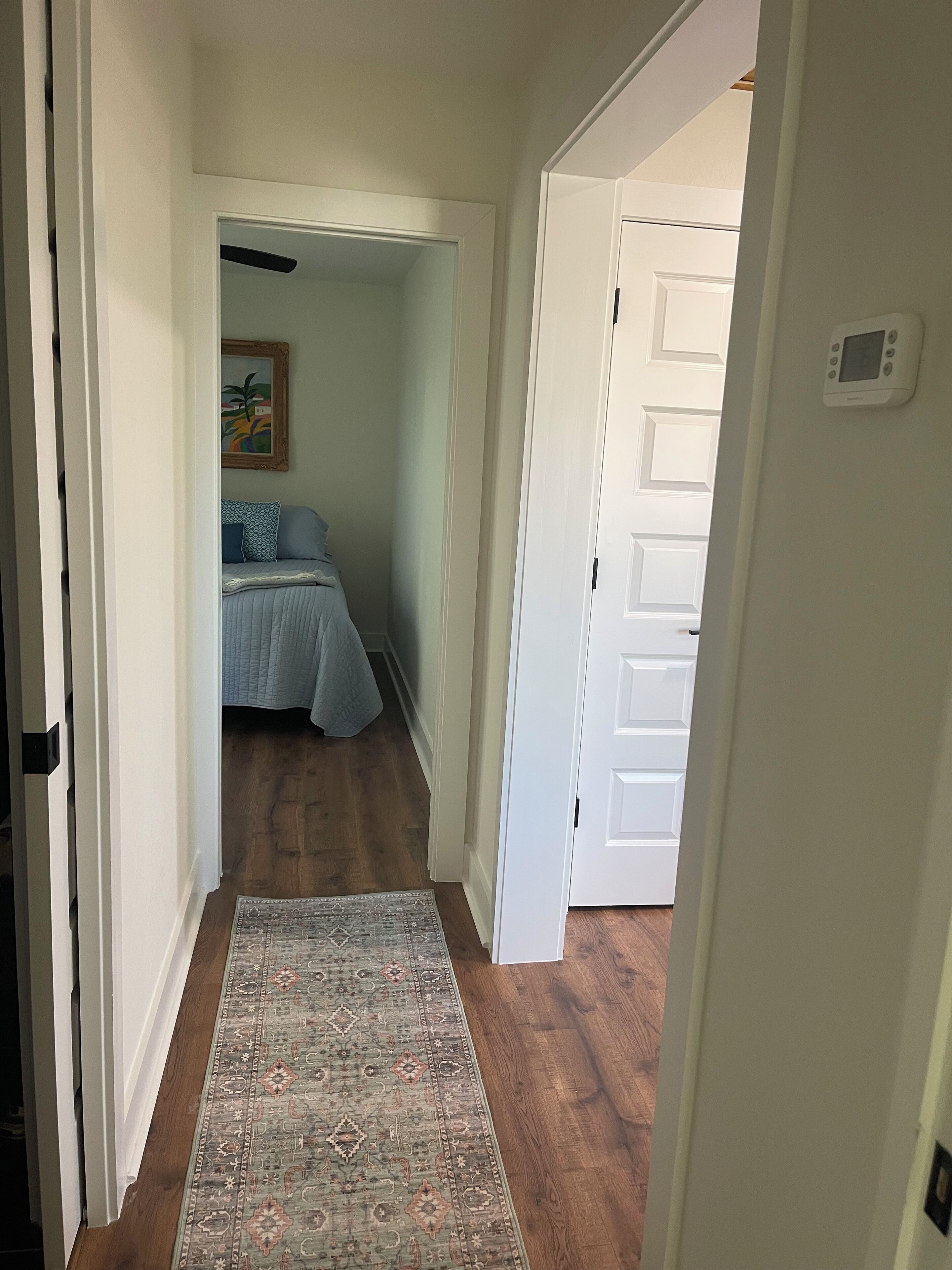 Un couloir étroit, orné d’un tapis à motifs, mène à une chambre à coucher où se trouvent un lit et un tableau accroché au mur.