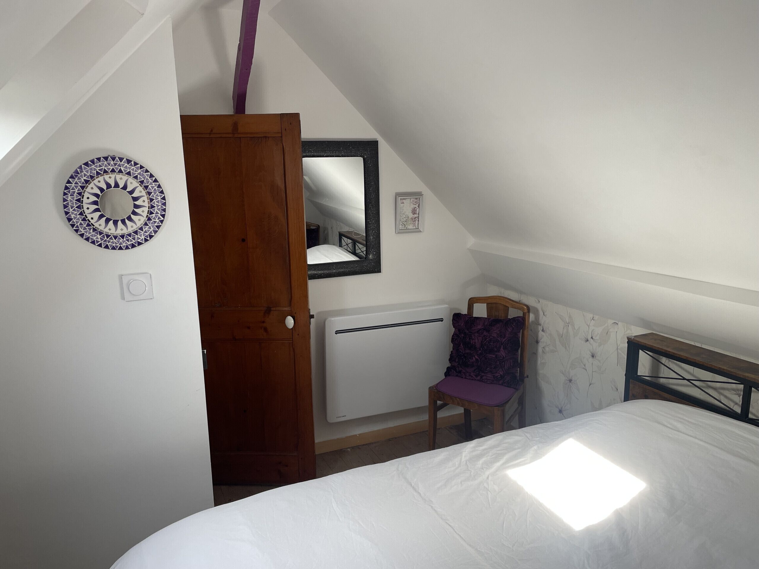 A bedroom with a bed, a chair, a wooden door, a mirror, and a wall-mounted radiator.