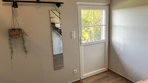 Ein Zimmer mit weißer Tür, einem Fenster mit Blick auf Grünflächen, einer hängenden Pflanze und einem Spiegel an der Wand.