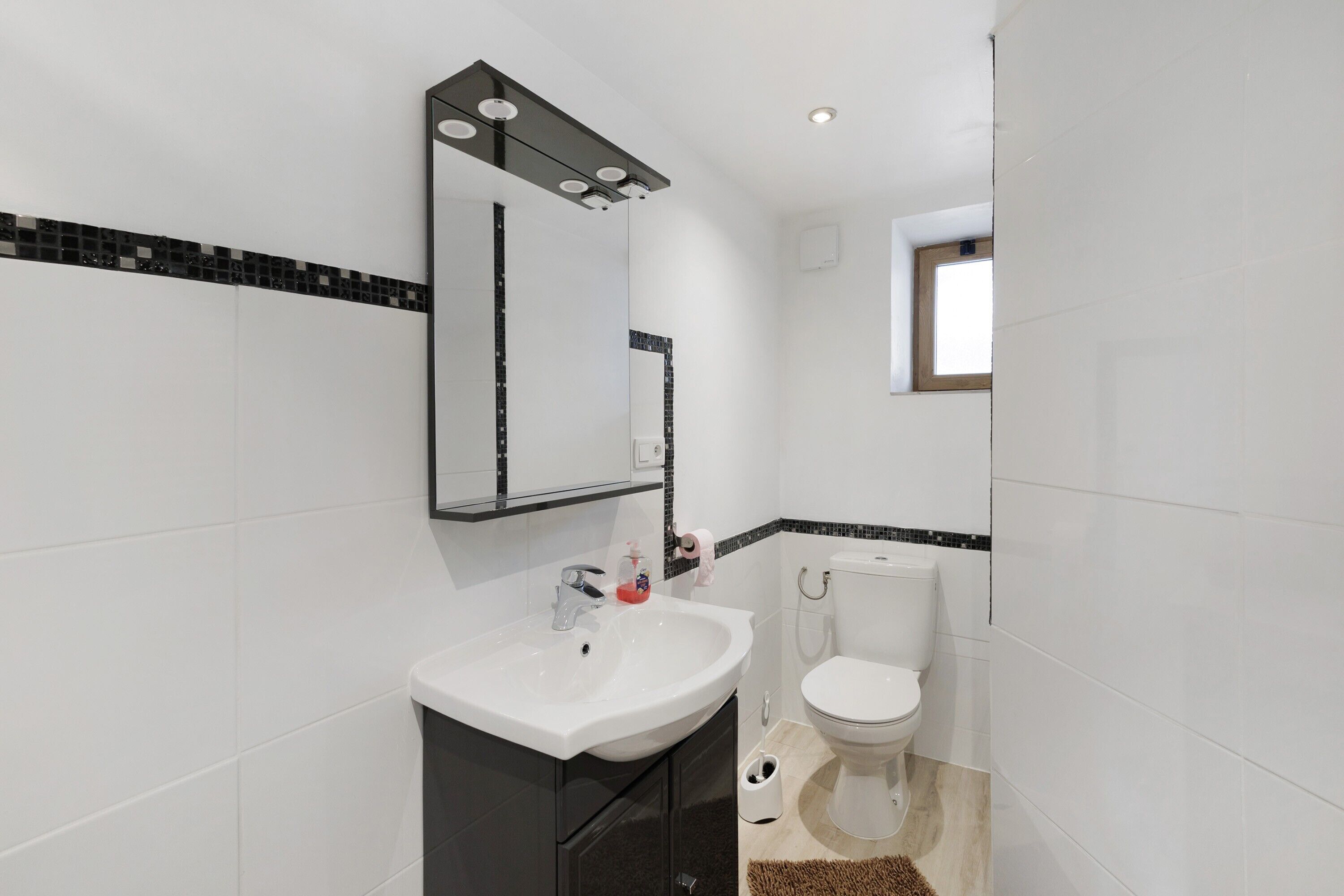 Un baño con un lavamanos blanco, un espejo con iluminación, un inodoro y una ventana.